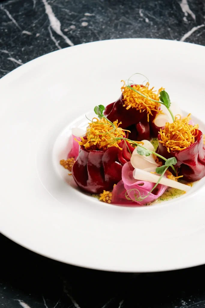 A white plate with an artfully arranged gourmet dish featuring rolled beet slices, crispy fried garnish, pickled vegetables, and microgreens on a dark marble surface at Alcron Restaurant.